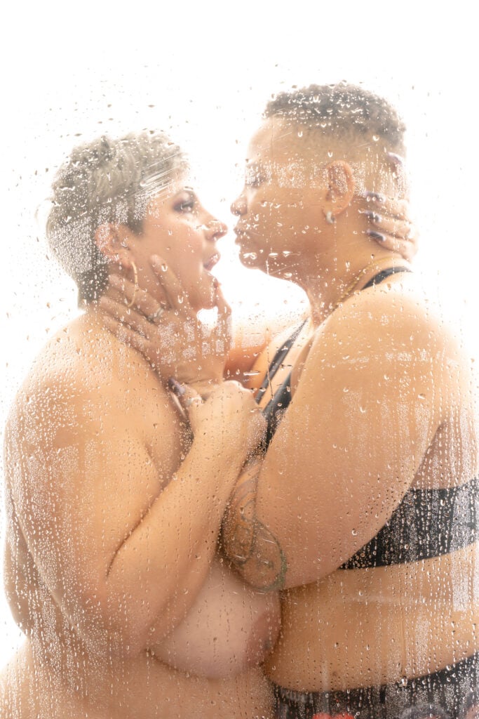 Couple in the shower, with a partner on the left compeltely naked just wearing dark makeup and a large gold hoop. Her partner is wearing a black sports bra. They hold each other close and look at each other longingly .Featured in the blog post "Femme vs Masc Boudoir Styling, Outfits, and Posing" colorado boudoir photography, Self love, Colorado boudoir photographer, gender exploration, gender neutrality, self-exploration, sex, gender, fashion, style, confidence, body image, be yourself, self-discover, self-discovery, comfortability, boudoir, boudoir posing, boudoir imagery, boudoir photo, inclusivity. Image by Embodied Art Boudoir.