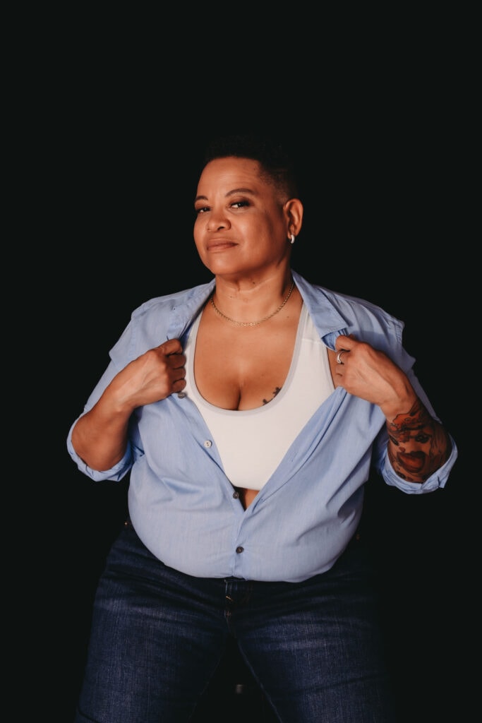 Black woman with a short buzz cut hair, wearing a blue button up shirt half-unbuttoned, exposing their white sports bra and silver chain necklace, with blue jeans. Featured in the blog post "Femme vs Masc Boudoir Styling, Outfits, and Posing" colorado boudoir photography, Self love, Colorado boudoir photographer, gender exploration, gender neutrality, self-exploration, sex, gender, fashion, style, confidence, body image, be yourself, self-discover, self-discovery, comfortability, boudoir, boudoir posing, boudoir imagery, boudoir photo, inclusivity. Photograph by Embodied Art Boudoir.