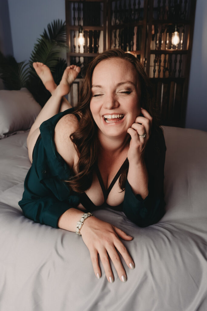 Brunette woman with wavy hair pushed to the side lying on a bed with grey sheets in a calming room smiling and laughing showcasing her white teeth, she is wearing a blue robe with a strappy bra and a bracelet. Featured in the blog "Why do a Boudoir Shoot?"Why, intentional photoshoot, intentions, experience, boudoir photography, confidence building, self-care, self-love, self-discover, personal growth, colorado boudoir, denver boudoir, boulder boudoir, colorado springs boudoir. Imagery by Embodied Art Boudoir.