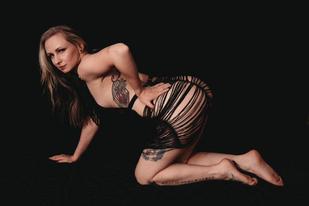 Female booty wearing a strappy dress, hunched over in front of a super dark backgrund and featuring her tattoo. Featured in the blog, "Why I Care So Much About Intention Setting…and Why It Matters For A Boudoir Photoshoot" ntentions, setting intentions, goals, listen to your heart, experience, growth, sexy, boudoir photo, boudoir images, self confidence, self love, personal tranformation. Image by Embodied Art Boudoir. 