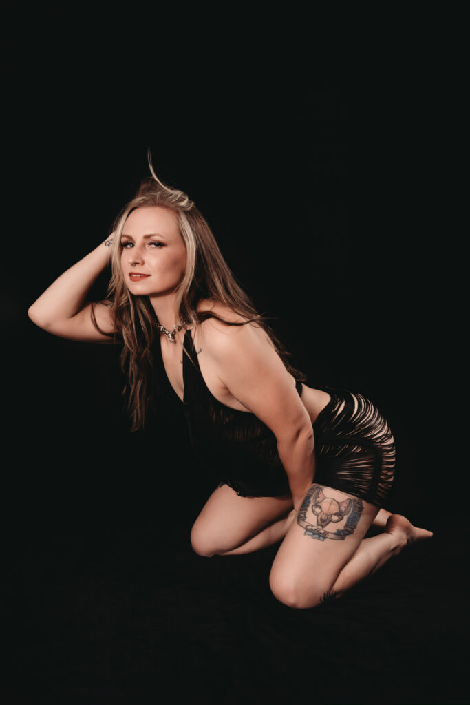 Blonde woman with thick highlights and straight, kneeling wearing a see through strappy black dress in front of a dark background, she looks seductively at the camera. Featured in the blog, "Why I Care So Much About Intention Setting…and Why It Matters For A Boudoir Photoshoot" ntentions, setting intentions, goals, listen to your heart, experience, growth, sexy, boudoir photo, boudoir images, self confidence, self love, personal tranformation. Image by Embodied Art Boudoir. 