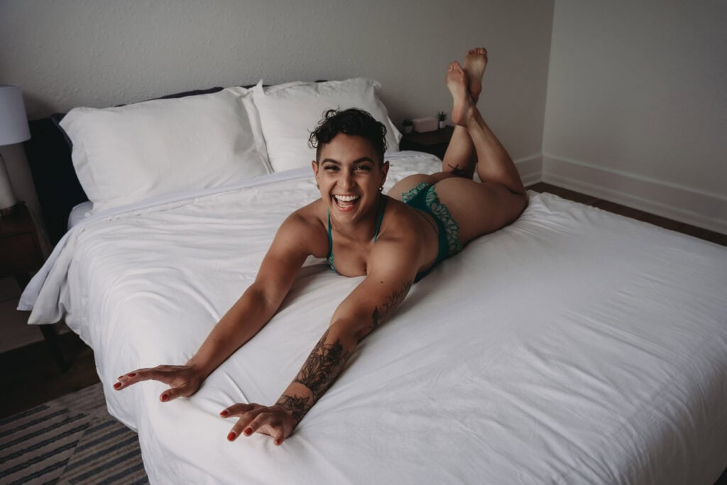 A woman with a brown complexion, a short curly tapered haircut, green strappy lingerie, red nail polish, and an abstract tattoo on her right arm is lying on a bed with white sheets. She is happily smiling while extending her arms forward and legs upward. Featured on About Your Denver Boudoir Photographer. Colorado boudoir photographer, Denver boudoir photographer, Denver boudoir photography, Colorado boudoir photography, self-love, mindfulness, body positivity, fun photo shoot