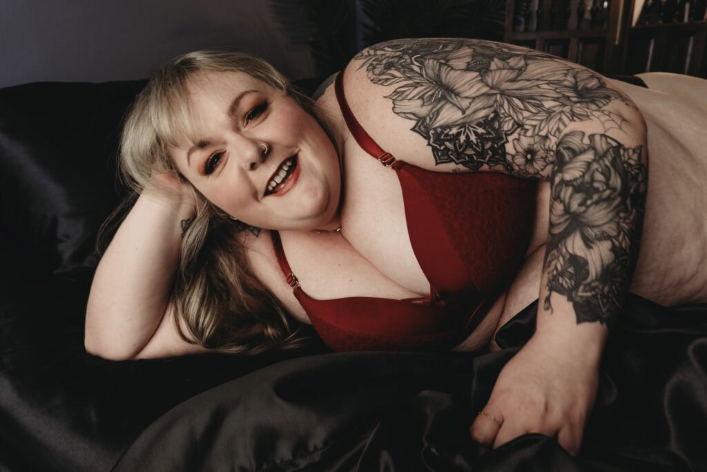 A pale, plus-sized woman with long blonde hair, peach makeup, a red bra, and nose piercings is lying on a bed with black silk sheets. She has a floral arm sleeve tattoo on her left arm. She is posed with her right hand supporting her head while smiling. Featured on Denver Tattoo Photoshoot. Colorado tattoo photographer, Denver tattoo photographer, Denver boudoir photographer, Denver boudoir photography, Colorado boudoir photographer, body positivity, self-love