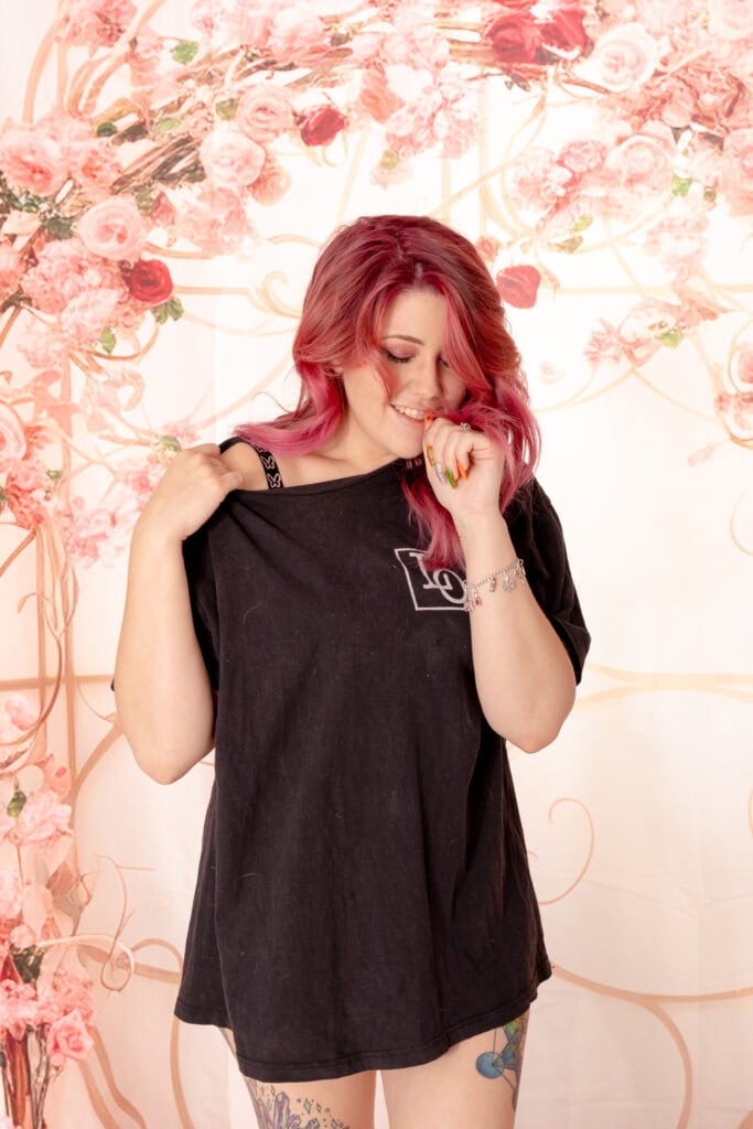 Woman with dyed pink hair standing in front of a floral pink background, wearing a oversized black t-shirt with a logo on the left breast, and her black bra strap is poking out. she is biting her nail and looking down, mysteriously. Featured in the blog "Boudoir Alibis: What to Tell Your Partner or Coworkers About Where You'll Be Next Tuesday" good excuse, alibi, secret, boudoir photoshoot, boudoir shoot, colorado boudoir, denver boudoir, golden colorado, surprise shoot, confidence, body journey, photo album. image by Embodied Art Boudoir.