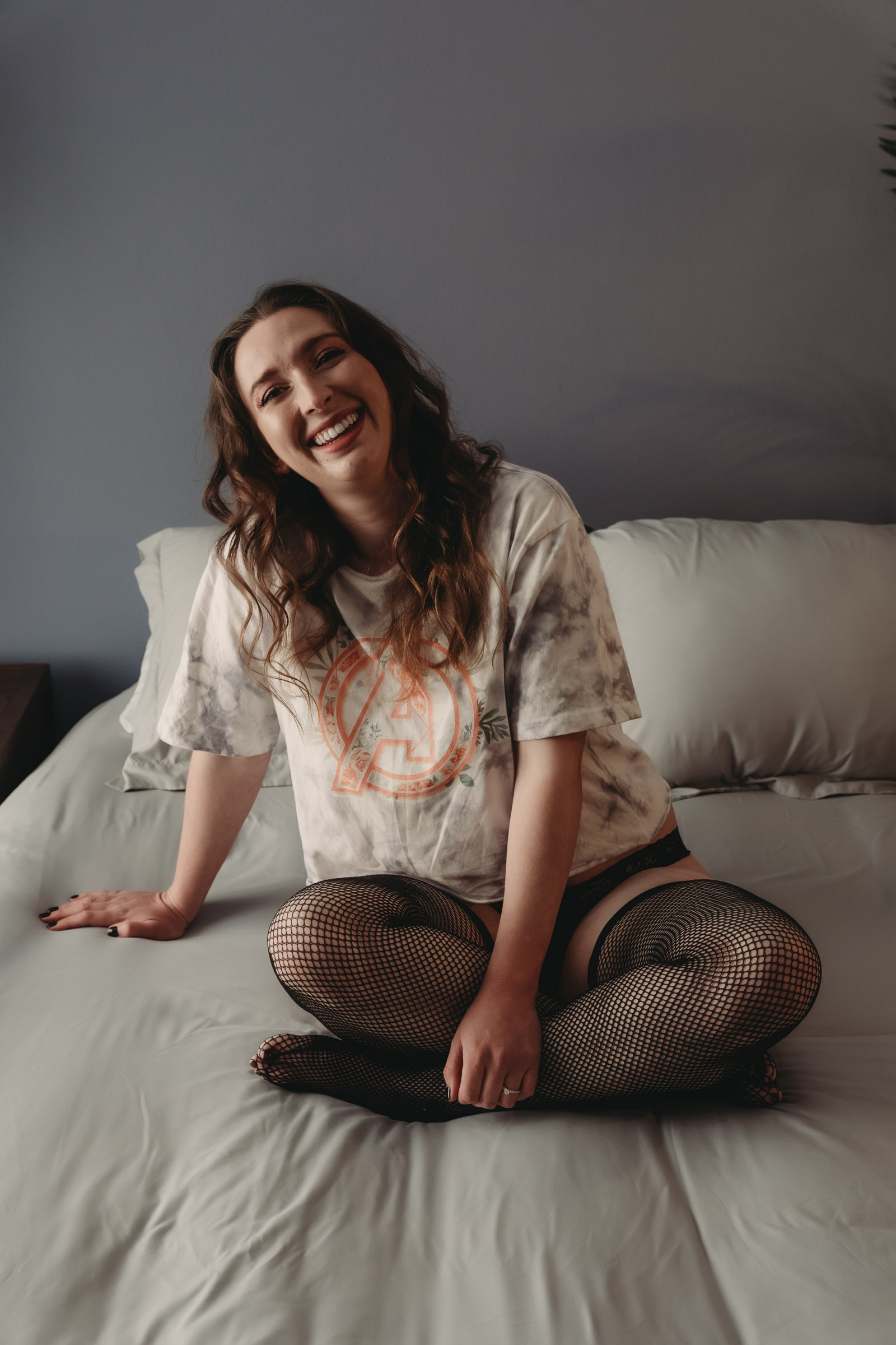 A woman with pale skin and long hair is sitting with her legs crossed on a bed with gray covers. She is wearing a white tie-dye shirt, black underwear, and fishnet stockings. Featured on Boudoir Sessions: Just the Basics. Photograph by Embodied Art Boudoir. Denver boudoir photographer, Denver boudoir photography, Colorado boudoir photographer, Colorado boudoir photography, boudoir photoshoot, mindfulness, body positivity, boudoir photography Denver