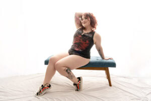 A pale woman with red curly hair is sitting on a blue bench. She has an arm and leg tattoo and is wearing a black graphic tank top, black lace underwear, and colorful sneakers. She is posed with her hand on her head and a confident expression on her face. Featured on Boudoir Sessions Treat Yourself. Image by Embodied Art Boudoir. Denver boudoir photographer, Denver boudoir photography, Colorado boudoir photographer, Colorado boudoir photography, body-positive boudoir, plus size boudoir, body image, boudoir photography Denver