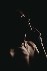 A bodyscape of a woman touching their lips, with soft lighting highlighting their face and upper body against a dark background. Featured on Denver Boudoir Photography - Body Positive Boudoir for 2025. Image by Embodied Art Boudoir. Denver boudoir photographer, Colorado boudoir photographer, Colorado boudoir photography, boudoir photoshoot, self-love, body confidence
