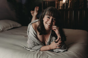A middle aged woman with dark and gray bangs and curled hair, wearing a striped robe, body chain, and unique stone rings, is joyfully smiling while lying on a bed. Featured on Denver Boudoir Photography - Body Positive Boudoir for 2025. Image by Embodied Art Boudoir. Denver boudoir photographer, Colorado boudoir photographer, Colorado boudoir photography, boudoir photoshoot, self-love, meditation