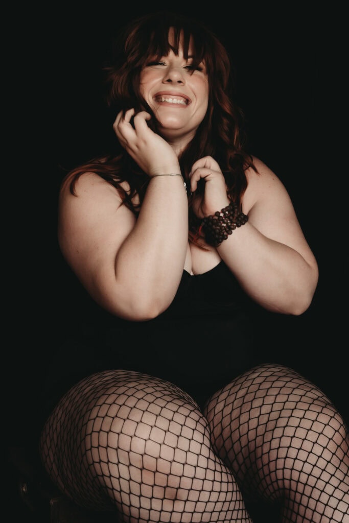 A woman with pale skin, long dark hair, and bangs, is blushing and smiling warmly, sitting against a dark background, wearing fishnet stockings, black bodysuit, and stacked beaded bracelets. Featured on Denver Boudoir Photography - Body Positive Boudoir for 2025. Photograph by Embodied Art Boudoir. Denver boudoir photographer, Colorado boudoir photographer, Colorado boudoir photography, boudoir photoshoot, body image, mindfulness