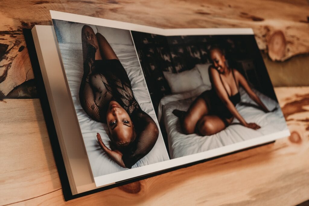 An open photobook is placed on a wooden surface, displaying images of a black woman posing on a bed, wearing a black outfit. Featured on Denver Boudoir Photography - Body Positive Boudoir for 2025. Photograph by Embodied Art Boudoir. Denver boudoir photographer, Colorado boudoir photographer, Colorado boudoir photography, boudoir photoshoot, empowerment, body image