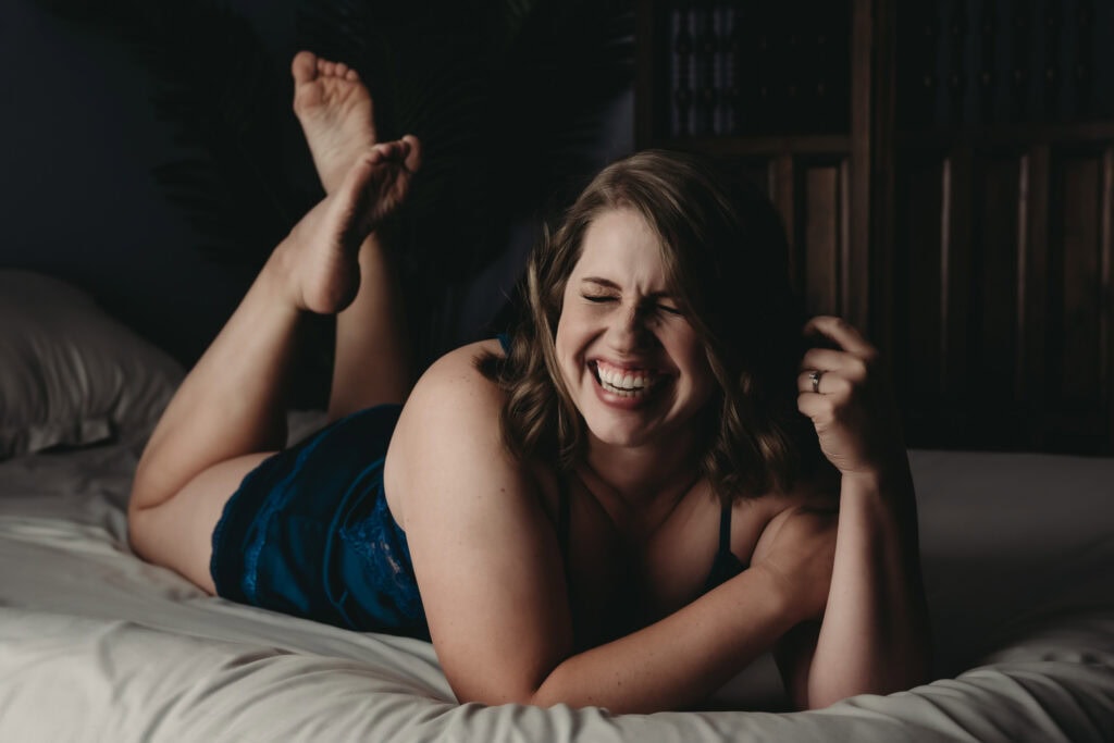 A woman with pale skin and light brown hair is lying on her stomach on a bed with gray covers, wearing a dark blue silk nightdress, smiling and laughing with eyes closed. Featured on Denver Boudoir Photography - Body Positive Boudoir for 2025. Photo by Embodied Art Boudoir. Denver boudoir photographer, Colorado boudoir photographer, Colorado boudoir photography, boudoir photoshoot, body positivity, mindfulness