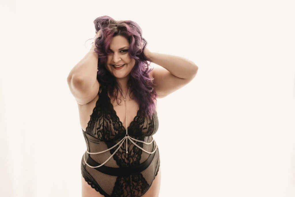 A woman with curly purple hair, wearing a black lace lingerie and a silver chain accessory is posed with her hands on her head and smiling in front of a white background. Featured on Denver Boudoir Photography - Body Positive Boudoir for 2025. Photograph by Embodied Art Boudoir. Denver boudoir photographer, Colorado boudoir photographer, Colorado boudoir photography, boudoir photoshoot, body image, body positivity