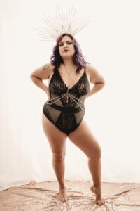 A woman with purple hair, wearing one-piece black lace lingerie, body chains, and a decorative hair piece stands confidently in a room with a sheer white curtain as the background. Featured on Denver Boudoir Photography - Body Positive Boudoir for 2025. Photograph by Embodied Art Boudoir. Denver boudoir photographer, Colorado boudoir photographer, Colorado boudoir photography, boudoir photoshoot, self-love, self confidence