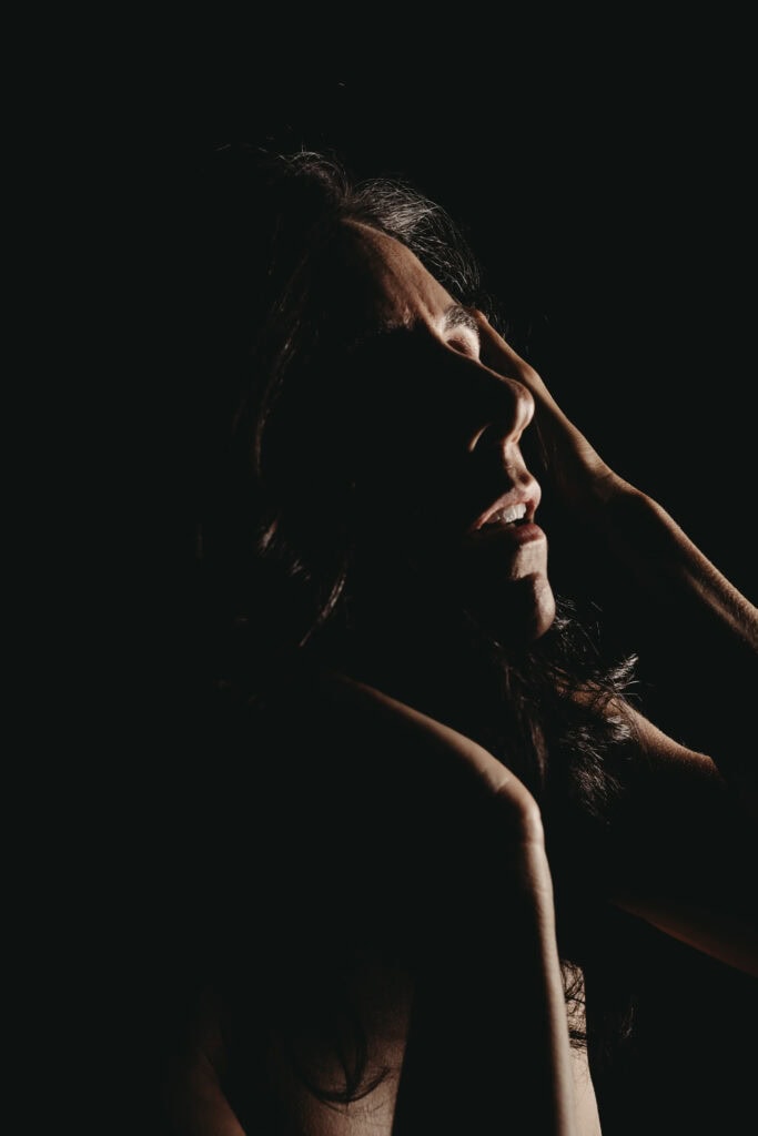 A bodyscape of a woman with curly, dark and gray hair, is posing and gently touching her face, illuminated by dramatic lighting against a dark background. Featured on Denver Boudoir Photography - Body Positive Boudoir for 2025. Photo by Embodied Art Boudoir. Denver boudoir photographer, Colorado boudoir photographer, Colorado boudoir photography, boudoir photoshoot, mindfulness, body image