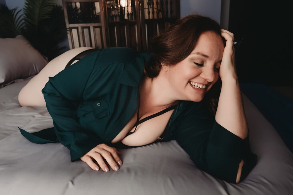 A woman with pale skin, dark hair, dark green long sleeves shirt, and black lingerie is lying on a bed, smiling with her eyes closed and her hand on her head. Featured on Denver Boudoir Photography - Body Positive Boudoir for 2025. Photograph by Embodied Art Boudoir. Denver boudoir photographer, Colorado boudoir photographer, Colorado boudoir photography, boudoir photoshoot, body confidence, body image