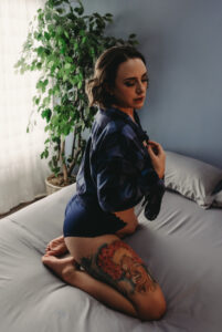 A woman with brown short hair, wearing blue checkered long sleeves and blue lingerie is kneeling on a bed while holding the collar of her shirt. She is looking down for a dramatic effect and has a colorful tattoo featuring a red tree on her right thigh. Featured on Denver Boudoir Photography - Body Positive Boudoir for 2025. Image by Embodied Art Boudoir. Denver boudoir photographer, Colorado boudoir photographer, Colorado boudoir photography, boudoir photoshoot, empowered, self-love