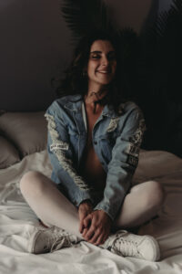 A woman sitting on a bed with white beddings, wearing a ripped denim jacket with patches, white stockings, and white shoes. Her eyes are closed, and they are smiling slightly, with their hands clasped. Featured on Denver Boudoir Photography - Body Positive Boudoir for 2025. Photograph by Embodied Art Boudoir. Denver boudoir photographer, Colorado boudoir photographer, Colorado boudoir photography, boudoir photoshoot, mindfulness, meditation