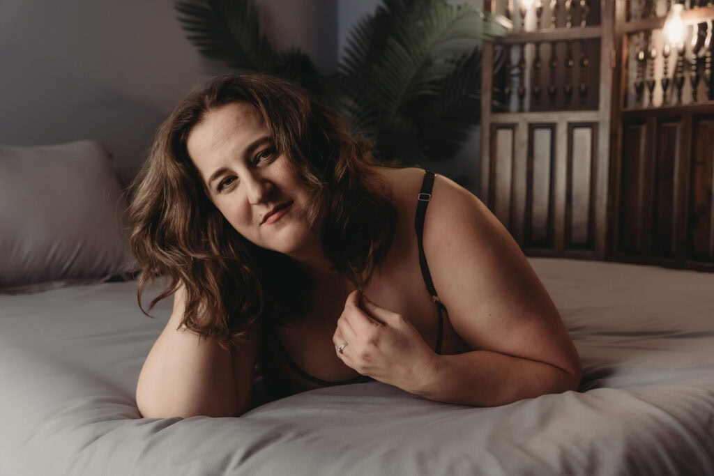 A woman with brown curly hair, wearing black lingerie is slightly smiling and lying on a bed with the bedroom having a cozy ambience. Featured on Denver Boudoir Photography - Body Positive Boudoir for 2025. Image by Embodied Art Boudoir. Denver boudoir photographer, Colorado boudoir photographer, Colorado boudoir photography, boudoir photoshoot, self-love, body image