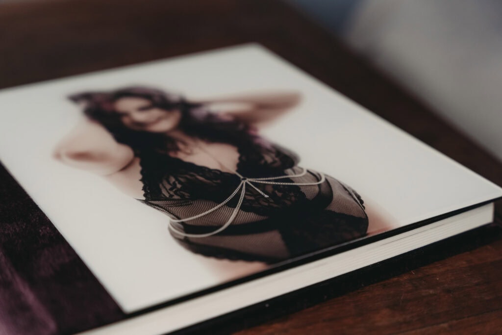 A woman wearing sexy black lingerie and a silver body chain is featured on the cover of the brown velvet photo album which is placed on top of a wooden table. Featured on Denver Boudoir Photography - Body Positive Boudoir for 2025. Photograph by Embodied Art Boudoir. Denver boudoir photographer, Colorado boudoir photographer, Colorado boudoir photography, boudoir photoshoot, plus size boudoir, body confidence