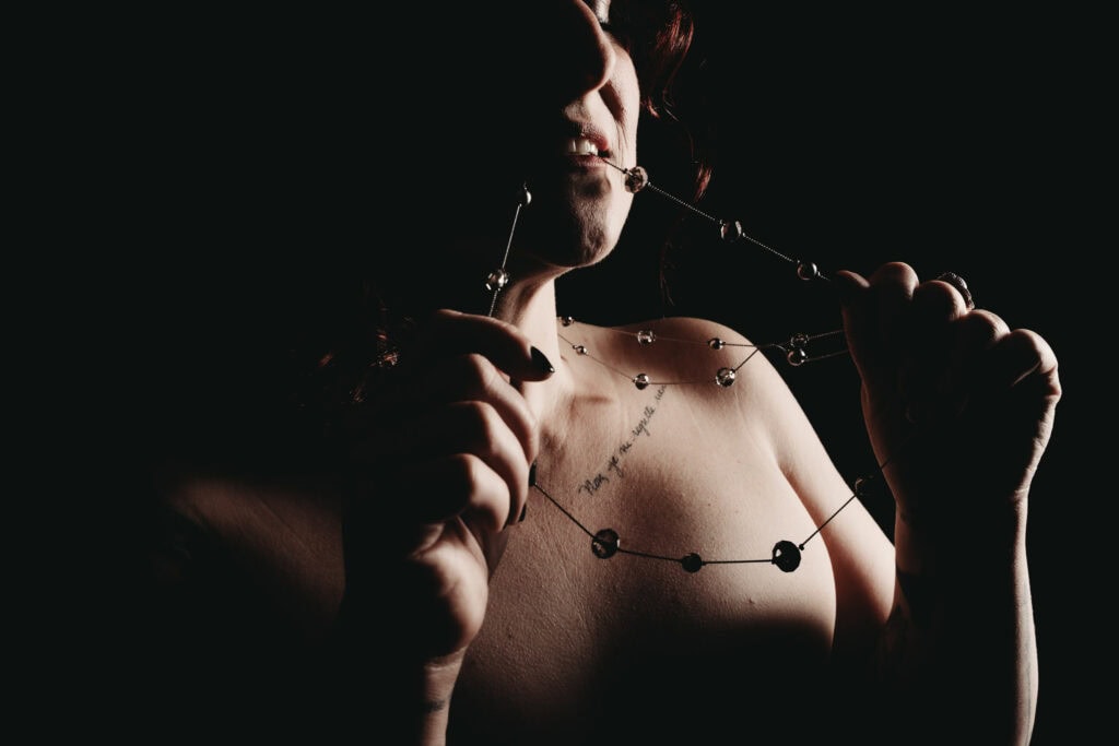A woman holding and slightly biting a beaded chain necklace, with a collar tattoo is posed under dramatic lighting, emphasizing her upper body. Featured on Denver Boudoir Photography - Body Positive Boudoir for 2025. Photo by Embodied Art Boudoir. Denver boudoir photographer, Colorado boudoir photographer, Colorado boudoir photography, boudoir photoshoot, empowerment, body image