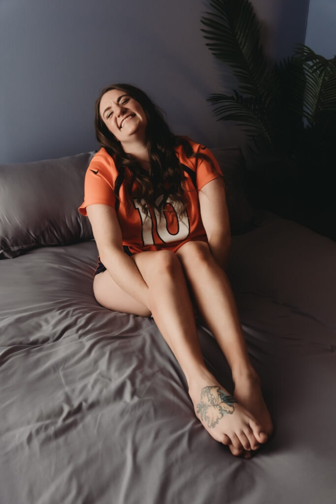 A woman with pale skin and long dark curly hair, a tattoo on her right foot, wearing an orange graphic shirt, is happily smiling and sitting on a bed. She is posed with her hands behind her legs and her feet overlapped. Featured on Denver Boudoir Photography - Body Positive Boudoir for 2025. Image by Embodied Art Boudoir. Denver boudoir photographer, Colorado boudoir photographer, Colorado boudoir photography, boudoir photoshoot, empowered, body confidence