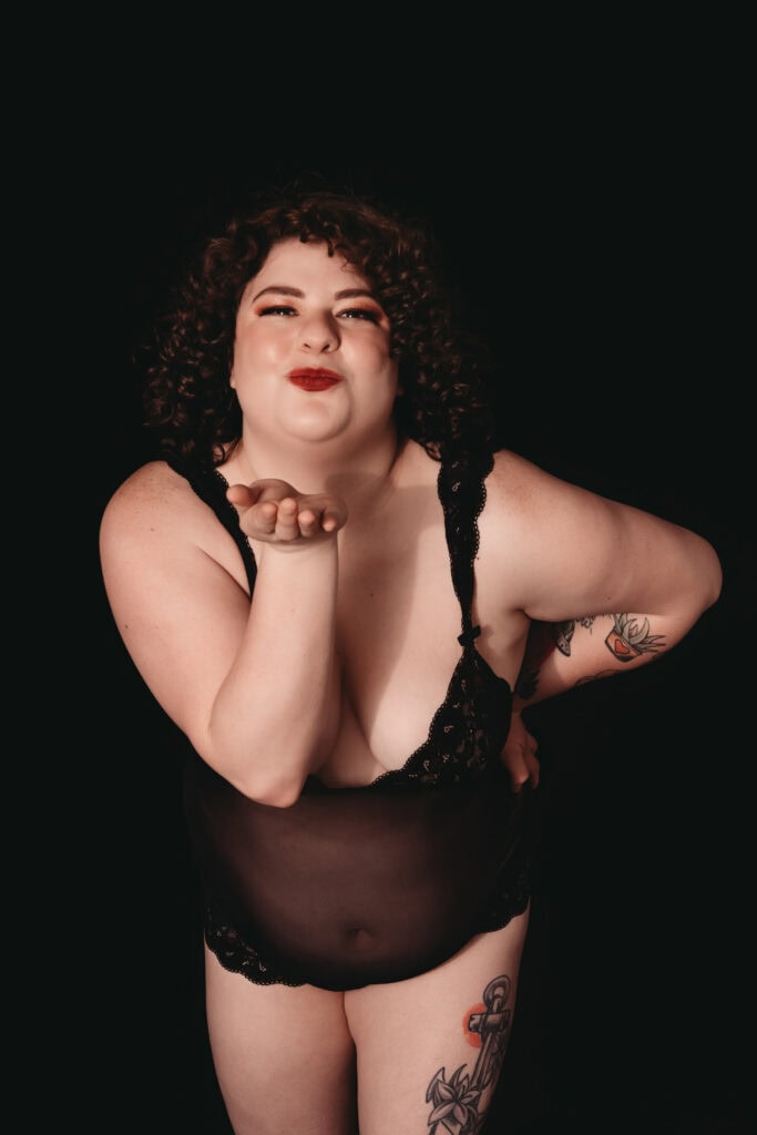 A plus size woman with curly hair blows a kiss while wearing black see-through lingerie. She has tattoos on her arm and thigh. Featured on Denver Boudoir Photography - Body Positive Boudoir for 2025. Photograph by Embodied Art Boudoir. Denver boudoir photographer, Colorado boudoir photographer, Colorado boudoir photography, boudoir photoshoot, plus size boudoir, body confidence