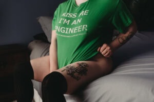 A woman with pale skin wearing a green t-shirt with the text saying "Kiss Me I'm an Engineer," is sitting on the edge of the bed while pulling her shirt down. She has black and colored tattoos on their thigh and arm, and is wearing black thigh-high socks. Featured on Denver Boudoir Photography - Body Positive Boudoir for 2025. Photograph by Embodied Art Boudoir. Denver boudoir photographer, Colorado boudoir photographer, Colorado boudoir photography, boudoir photoshoot, empowered, body confidence