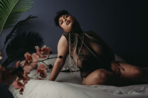 A woman with short curly black hair wearing a black mesh outfit and body chains poses on a bed with gray covers, with a backdrop of green leaves and pink flowers in the foreground. Featured on Denver Boudoir Photography - Body Positive Boudoir for 2025. Photo by Embodied Art Boudoir. Denver boudoir photographer, Colorado boudoir photographer, Colorado boudoir photography, boudoir photoshoot, self-love, plus size boudoir