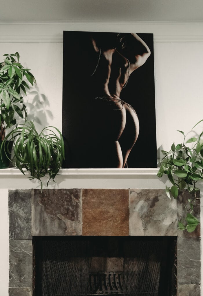 A printed bodyscape artwork with dramatic lighting featuring a muscular back and torso of a woman, draped with body chains, is placed above a fireplace with plants on either side. Featured on Denver Boudoir Photography - Body Positive Boudoir for 2025. Photograph by Embodied Art Boudoir. Denver boudoir photographer, Colorado boudoir photographer, Colorado boudoir photography, boudoir photoshoot, body image, body confidence