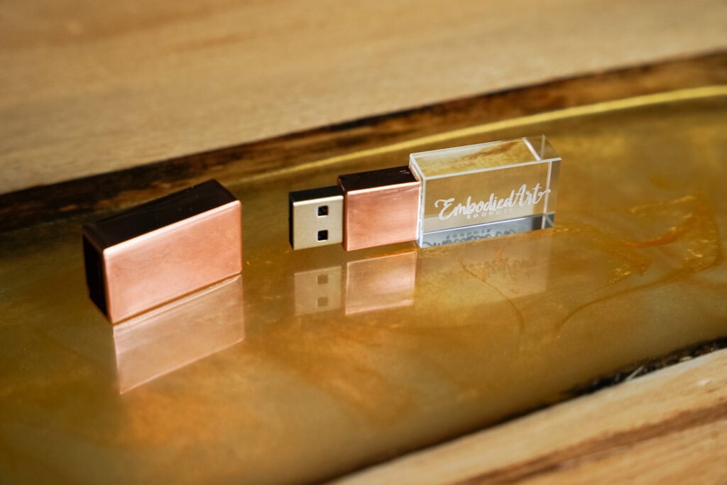 A USB flash drive with a copper cap and a clear casing displaying text “Embodied Art” is placed on a reflective golden surface. Featured on Denver Boudoir Photography - Body Positive Boudoir for 2025. Image by Embodied Art Boudoir. Denver boudoir photographer, Colorado boudoir photographer, Colorado boudoir photography, boudoir photoshoot, mindfulness, body image