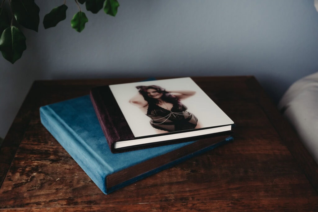 Two hardcover photo albums are stacked on a wooden bedside table next to a leaf decoration. A woman wearing sexy black lingerie and a silver body chain is featured on the cover of the brown velvet photo album. Featured on Denver Boudoir Photography - Body Positive Boudoir for 2025. Image by Embodied Art Boudoir. Denver boudoir photographer, Colorado boudoir photographer, Colorado boudoir photography, boudoir photoshoot, gift ideas, meditation
