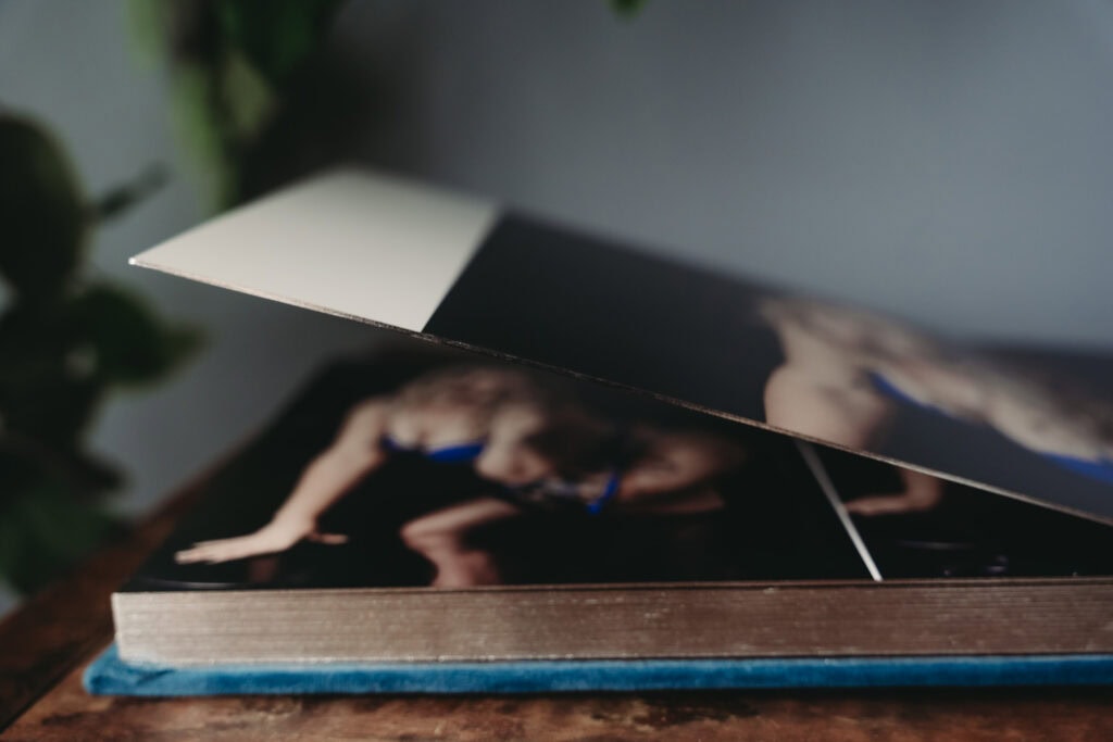 The image features the lower side of an open photo album on a wooden table displaying an image of a woman in blue lingerie, posing against a dark background. Featured on Denver Boudoir Photography - Body Positive Boudoir for 2025. Photograph by Embodied Art Boudoir. Denver boudoir photographer, Colorado boudoir photographer, Colorado boudoir photography, boudoir photoshoot, self-love, mindfulness