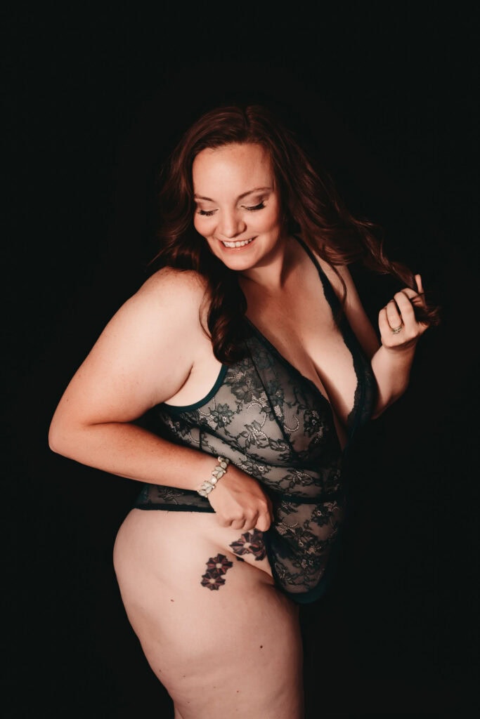 Tall woman with auburn curled hair smiling at the camera but looking away, wearing a low cut lacy flowery black body suit showcasing her floral tattoos on her leg and stomach. She is standing in front of a black background. Featured in the blog "Embracing Body Positivity Through Breathwork and Bodywork: A Journey at Bloom & Breath Bodywork" body positivity, body work, breathwork, mindful, mindfulness, bodywork, meditation, breathe, connection, body mind connection, healing, empowerment, calm, unlock healing, natural healing, holistic journey, denver colorado, denver co, breathing practices, calm the nervous system, self love, affirmation, heal. Picture by Embodied Art Boudoir.