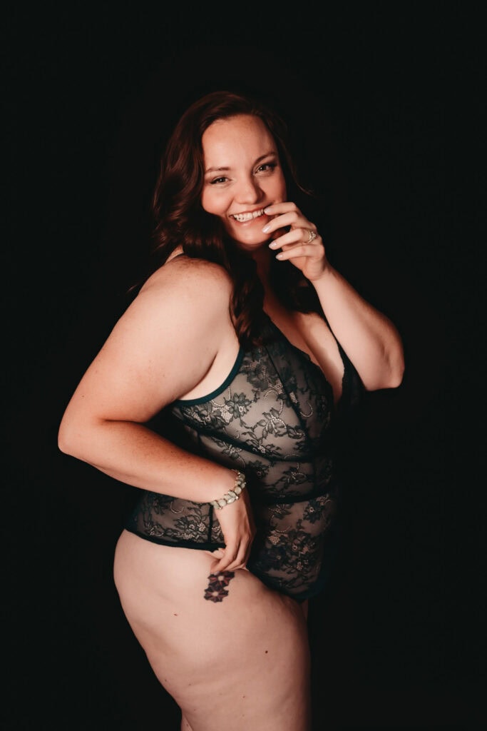 Tall woman with auburn curled hair smiling at the camera biting her finger sassily, wearing a low cut lacy flowery black body suit showcasing her floral tattoos on her leg and stomach. She is standing in front of a black background. Featured in the blog "Embracing Body Positivity Through Breathwork and Bodywork: A Journey at Bloom & Breath Bodywork" body positivity, body work, breathwork, mindful, mindfulness, bodywork, meditation, breathe, connection, body mind connection, healing, empowerment, calm, unlock healing, natural healing, holistic journey, denver colorado, denver co, breathing practices, calm the nervous system, self love, affirmation, heal. Photo by Embodied Art Boudoir.