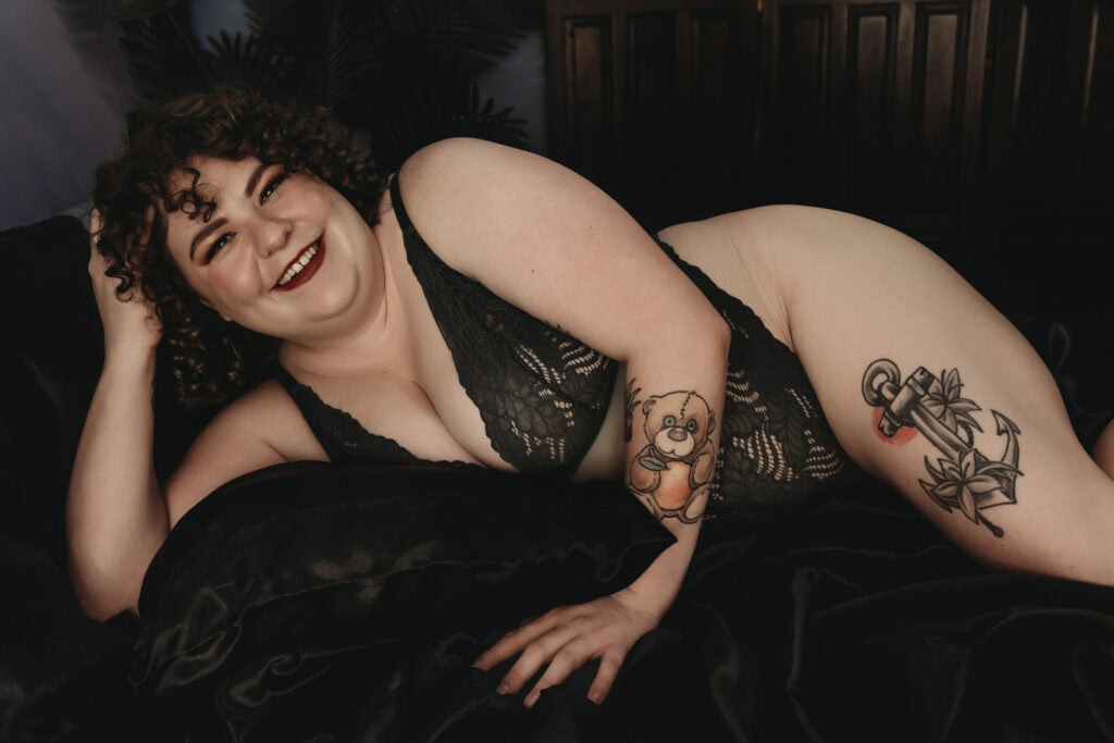 A pale, plus-size woman with short curly hair in black lace lingerie, and classic glam makeup with bold red lipstick, is lying on a bed with black silk sheets. She has tattoos of a colored teddy bear and plant on her arm and an anchor with flowers on her thighs. Featured on The Boudoir Lingerie Hub. Photo by Embodied Art Boudoir. Denver boudoir photographer, Denver boudoir photography, Colorado boudoir photographer, Colorado boudoir photography, plus size boudoir, boudoir photoshoot, body confidence
