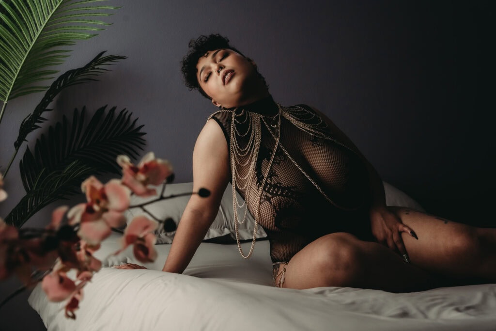 A woman with short curly black hair wearing a black mesh body suit and body chains poses with her eyes closed on a bed with gray sheets. The bedroom has a backdrop of gray walls, green leaves, and pink flowers in the foreground. Featured on The Boudoir Lingerie Hub. Photograph by Embodied Art Boudoir. Denver boudoir photographer, Denver boudoir photography, Colorado boudoir photographer, Colorado boudoir photography, plus size boudoir, body confidence, self-love
