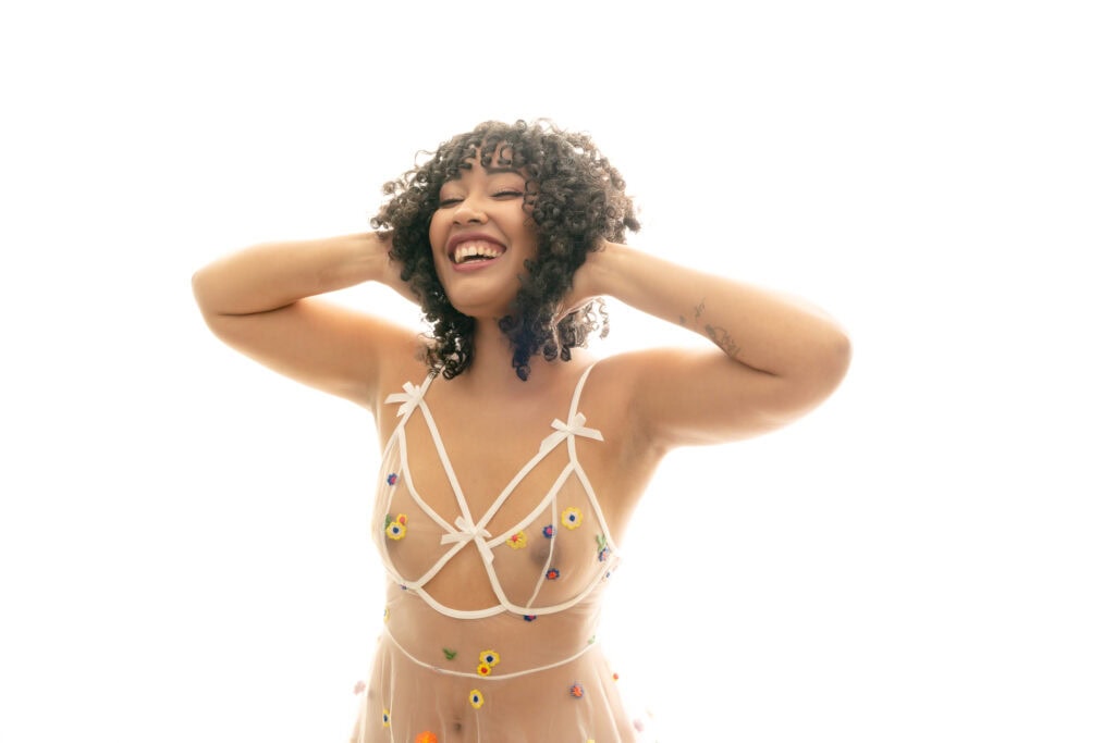 A woman with short curly hair, wearing sheer white lingerie with small colorful flowers, is smiling joyfully and posing with her hands on her hair against an overexposed white background. Featured on The Boudoir Lingerie Hub. Photo by Embodied Art Boudoir. Denver boudoir photographer, Denver boudoir photography, Colorado boudoir photographer, Colorado boudoir photography, meditation, mindfulness, self-love