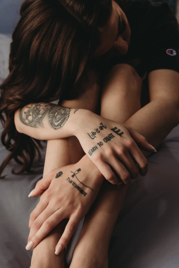 A woman with dark curly hair and black shirt is sitting on a bed with her arms crossed over her knees, focusing on the tattoos on her arms and hands. Featured on Denver Boudoir Photography - Body Positive Boudoir for 2025. Photograph by Embodied Art Boudoir. Denver boudoir photographer, Colorado boudoir photographer, Colorado boudoir photography, boudoir photoshoot, meditation, body image