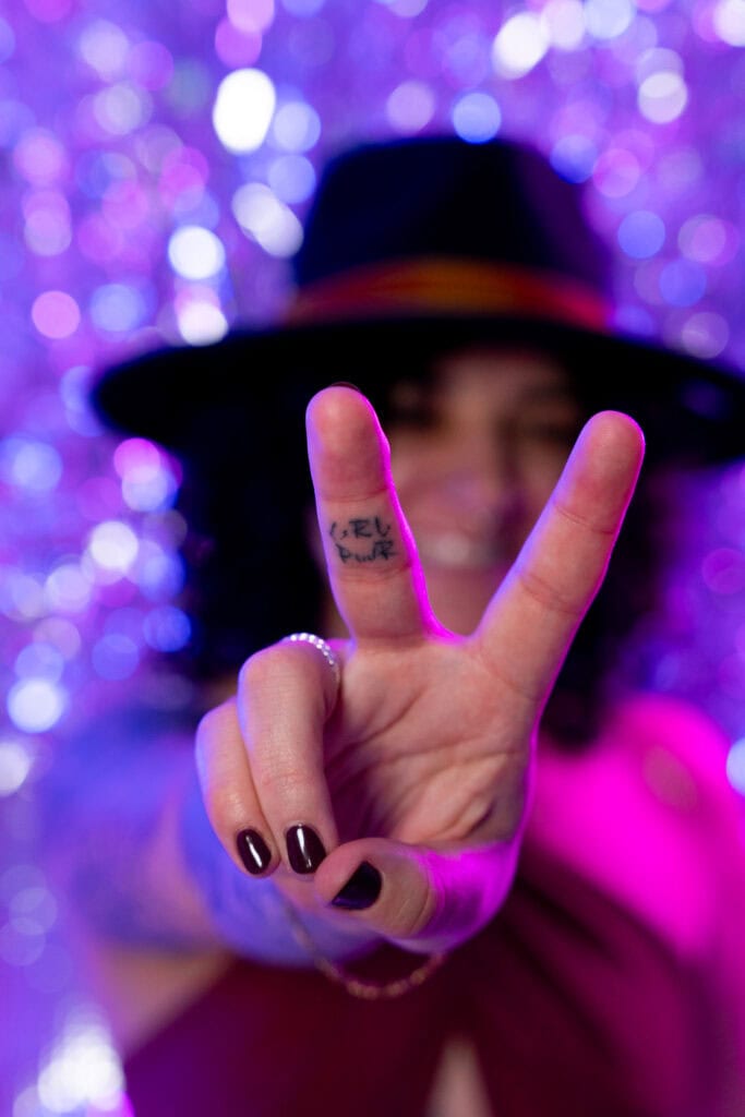 A woman wearing a black hat holds up her hand in a peace sign gesture, with the words "GRL PWR" tattooed on her fingers. She has dark nail polish and is wearing a ring and a bracelet. The background is blurred with purple and blue bokeh lights. Featured on Denver Boudoir Photography - Body Positive Boudoir for 2025. Image by Embodied Art Boudoir. Denver boudoir photographer, Colorado boudoir photographer, Colorado boudoir photography, boudoir photoshoot, body confidence, self-love