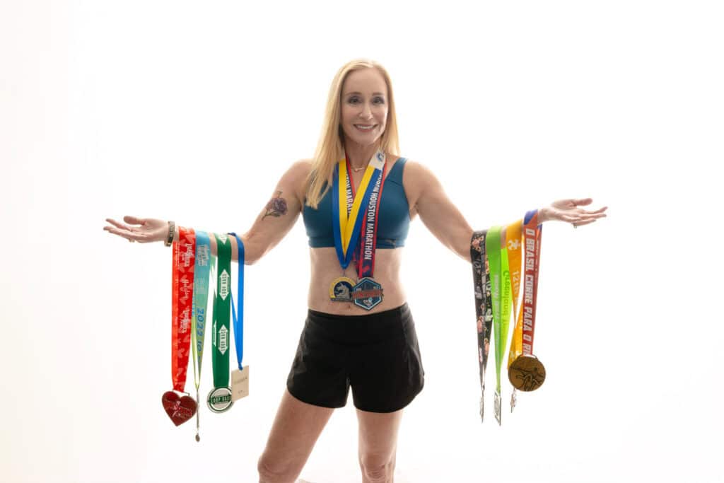 A woman with blonde hair and arm tattoo wearing an athletic outfit smiles as she displays several medals around her neck and arms, against a white background. Featured on Denver Boudoir Photography - Body Positive Boudoir for 2025. Photograph by Embodied Art Boudoir. Denver boudoir photographer, Colorado boudoir photographer, Colorado boudoir photography, boudoir photoshoot, empowerment, mindfulness