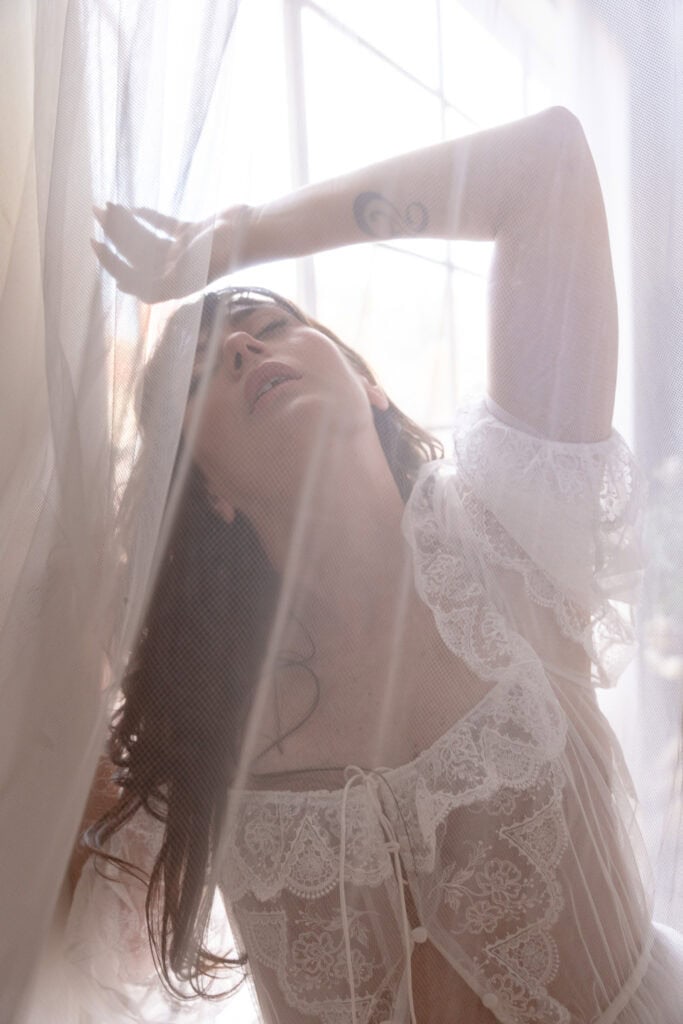 Woman with long brown hair moving gracefully around white lacy blinds, and she is wearing a lacy white t-shirt with designs. Featured in the blog, "Busting Excuses" excuses, bust excuse, myth, boudoir photoshoot, boudoir, love yourself, self-care, love your body, self-love, self-worth, validity, feeling good, your moment, posing, invest in yourself, boudoir photographer, female photographer, milestone, deserving, celebration, boudoir denver, Denver co. Imagery by Embodied Art Boudoir.