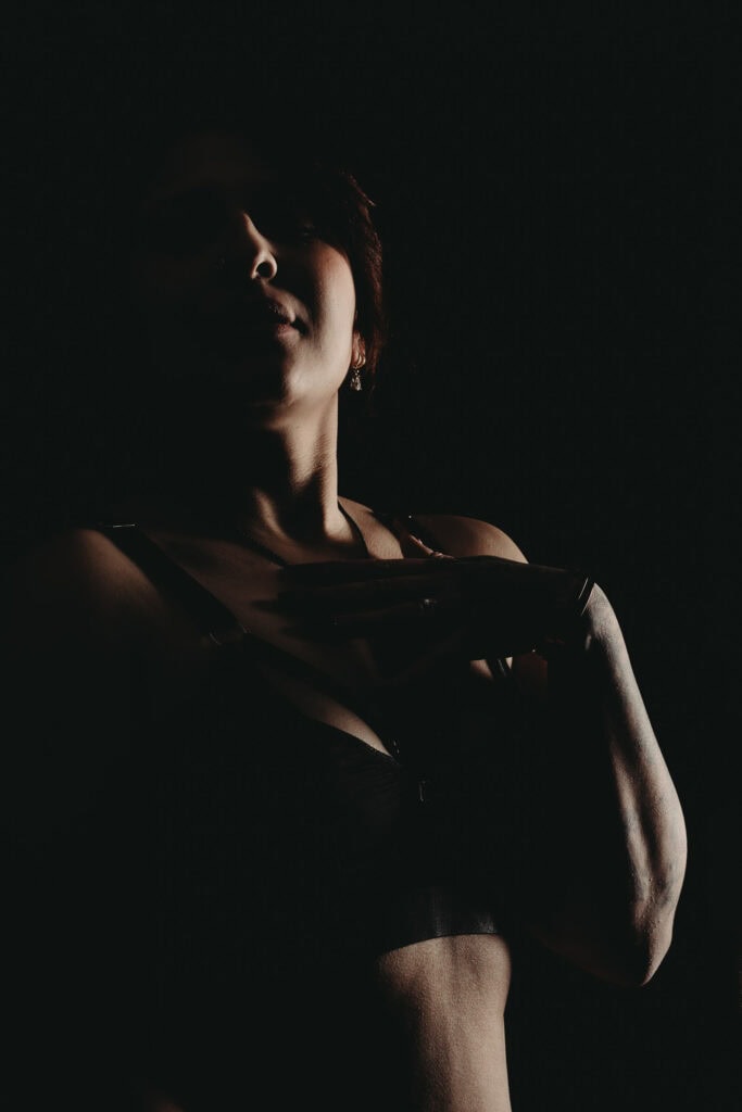 Silhouette of a woman wearing a black bra posing in front of a black background. Featured in the blog, "How Trust And Confidence Fuel Your Empowering Boudoir Experience." empowering boudoir photoshoot experience, boudoir photography for self-confidence, how to feel confident in a boudoir shoot, trust and vulnerability in boudoir, boudoir session for personal empowerment, feminine energy in boudoir photography, confidence-building photoshoot ideas, self-love through boudoir photography, celebrating your body with boudoir, transformational boudoir experience, healing through boudoir photography. Photo by Embodied Art Boudoir.