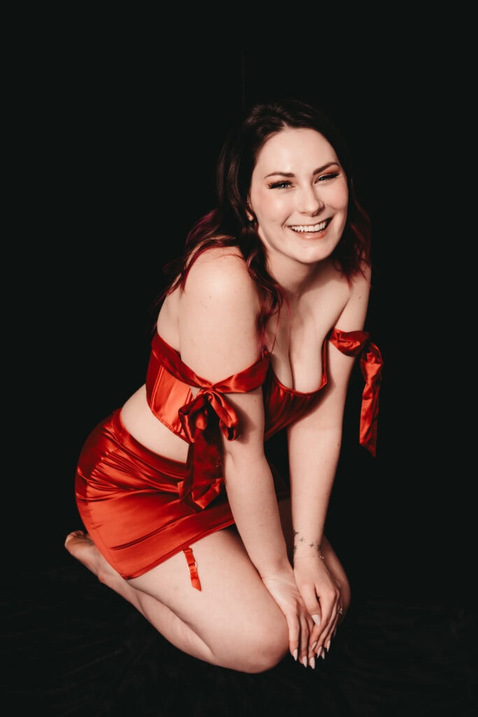 Brunette woman with wavy hair posing in front of a black background wearing a shiny red skirt and matching top with bows on the shoulders, smiling brightly at the camera with pink eyeshadow. Featured in the blog. "Boudoir Confidence Is Not A Prerequisite, It's The Result." boudoir confidence journey, self love photo shoot, empowering boudoir photography, luxury boudoir experience, confidence building photo session, body positive photography, self discovery through boudoir, golden colorado boudoir photographer, transformative photo shoot for women, embodied boudoir session, healing through photography, self empowerment photography, intimate portrait session, boudoir for self growth, mindful boudoir experience, boudoir for the soul, emotional boudoir session, inner goddess photo shoot, women’s self worth journey, authentic boudoir images. Photography by Embodied Art Boudoir.
