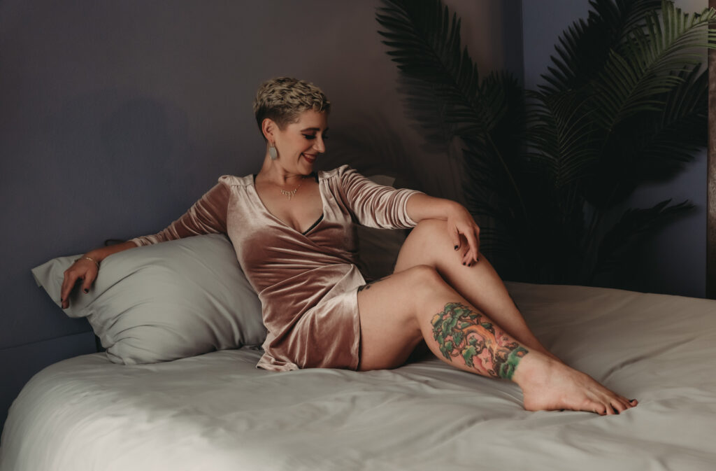 Blonde woman with short hair posing on a bed with crisp grey sheets, wearing a velvet baby pink drink with long sleeves and a plunging neckline, with gorgeous earrings and dark dramatic eye-makeup. Featured in the blog post, "Boudoir Photography Experience: A Day of Pampering, Self-Love, and Appreciation." Boudoir photography experience, self-love photo session, Colorado boudoir studio, women’s empowerment photography, confidence photoshoot, pampering day, luxury photo session, natural beauty portraits, minimal retouching, celebrating your body, emotive portraiture, personal transformation, feminine strength, authentic beauty. Photo by Embodied Art Boudoir.