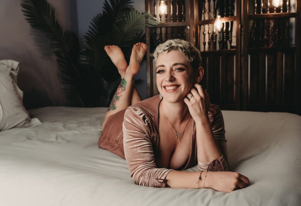 Blonde woman with short hair posing on a bed with crisp grey sheets, smiling beautifully at the camera with dramatic eye makeup and wearing a baby pink velvet dress with sleeves. Featured in the blog post, "Boudoir Photography Experience: A Day of Pampering, Self-Love, and Appreciation." Boudoir photography experience, self-love photo session, Colorado boudoir studio, women’s empowerment photography, confidence photoshoot, pampering day, luxury photo session, natural beauty portraits, minimal retouching, celebrating your body, emotive portraiture, personal transformation, feminine strength, authentic beauty. Photography by Embodied Art Boudoir.