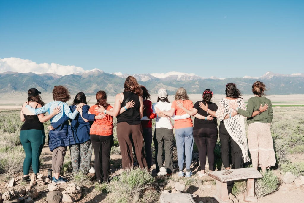 Group of 11 women standing with their back to the camera, arms around eachother, in casual, colorful clothing outside in front of a stunning large mountain range with plenty of foliage and grass in the foreground. Featured in the blog, "The Power & Importance Of Friendship." friendship, connection, community, come together, lonely, lonely women, seeking connection, friends, confidant, fun times, hardships, support, powerful, togetherness, beat loneliness, connection, belong, affection, intentional, beautiful. Photo by Embodied Art Boudoir.