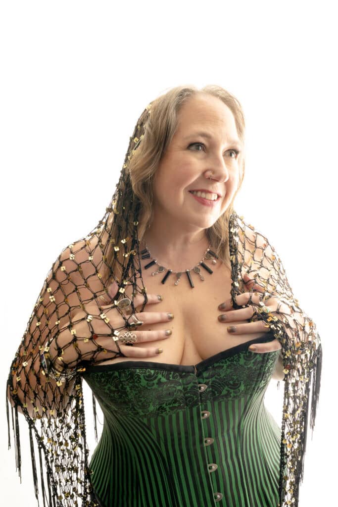 Mature woman with greying hair straightened wearing light makeup and a green and black lined corset with gold buttons and a black shawl with holes, and a statement necklace around her neck, smiling to the side. Featured in the blog, "The Power & Importance Of Friendship." friendship, connection, community, come together, lonely, lonely women, seeking connection, friends, confidant, fun times, hardships, support, powerful, togetherness, beat loneliness, connection, belong, affection, intentional, beautiful. Photograph by Embodied Art Boudoir.