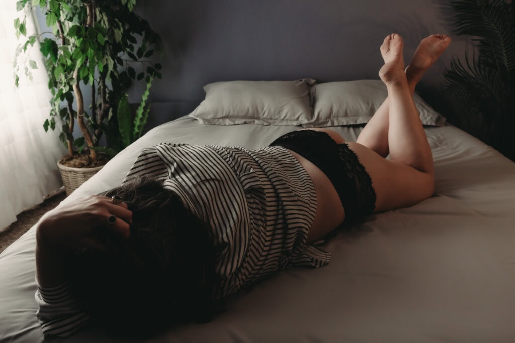 Woman lying on a bed with crisp white sheets wearing a striped black and white blouse with black full brief panties. Featured in the blog, "From Nervous To Thrilled: Rediscovering Beauty After A Lumpectomy." body positive boudoir photography, boudoir photography for self confidence, empowering boudoir session, self love photo shoot, celebrate your body boudoir, confidence boosting photography, boudoir experience for women, body acceptance photography, boudoir session for self esteem, see yourself differently photoshoot, self empowerment boudoir, women’s portrait photography, luxury boudoir experience, transformational boudoir photos, boudoir photography to love yourself. Image by Embodied Art Boudoir