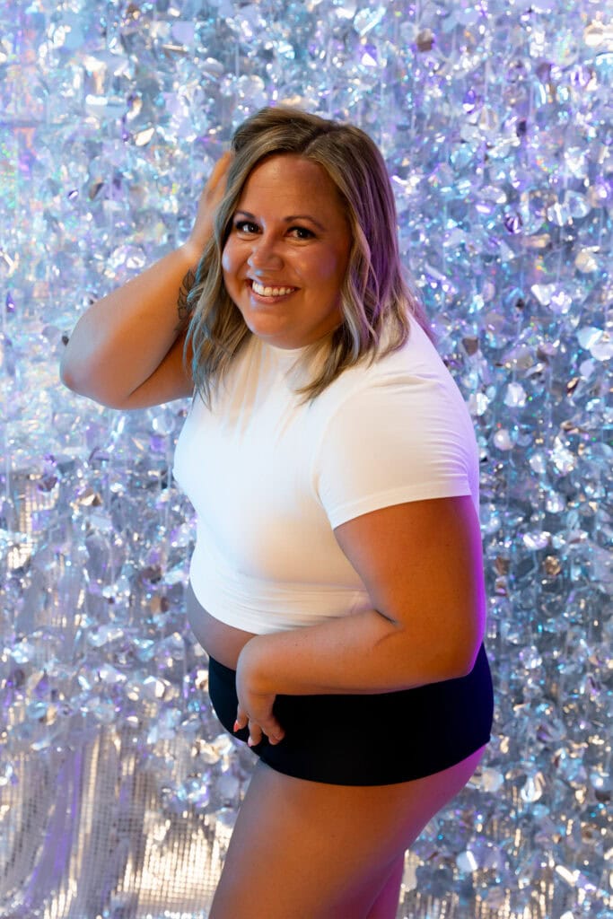 Brunette woman smiling in front of a disco background wearing a cropped tight white shirt with black shorts and smiling widely. Plus sized woman sitting on a blue bench wearing a black bodysuit smiling widely as she throws her head back gleefully. Featured in the blog, "Client Boudoir Experience: Joy, Play, and Empowerment in Every Frame." boudoir photography golden colorado, embodied art boudoir, gabby boudoir photographer, colorado boudoir studio, luxury boudoir experience, transformational boudoir session, self love boudoir, empowering boudoir photoshoot, women’s self love journey, confidence photography, body positive boudoir, mindful boudoir experience, feminine empowerment photography, self discovery photoshoot, artistic boudoir portraits, golden colorado photographer, denver boudoir photographer, soulful boudoir session, healing through photography, celebrate your body, authentic boudoir experience, embodied art experience, introspective boudoir, emotional healing photography, sensual self portrait, empowerment photography for women, boudoir photo inspiration, self confidence photoshoot, luxury women’s photography, intimate portrait experience. Photography by Embodied Art Boudoir.