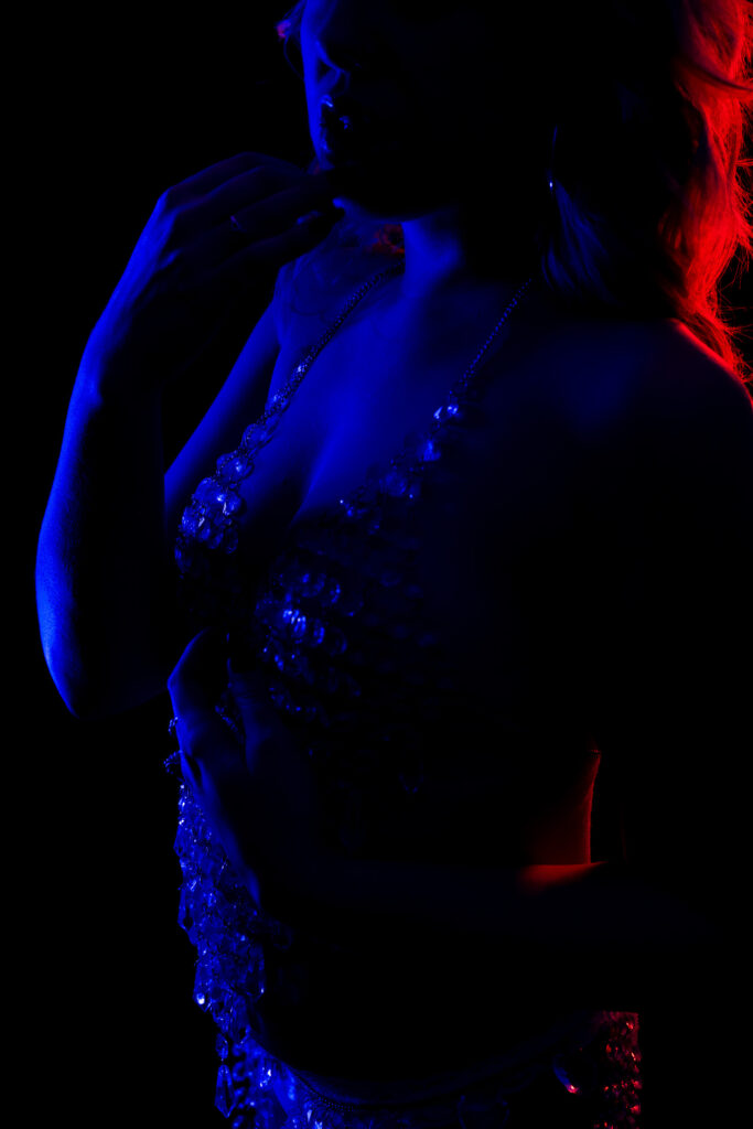 Close up of a woman posing in front of a black background, wearing a crystal bodysuit, holding her lip seductively. Featured in the blog "A Peek Behind the Curtain: The Creative Photoshoots That Feed My Soul." creative photography, creative photoshoot, artistic photography, experimental photography, fine art photography, creative portrait photography, body painting photography, artistic body paint, creative lighting photography, color lighting photography, studio portrait photography, personal photography project, behind the scenes photography, artistic self expression, Colorado photographer. Photos by Embodied Art Boudoir